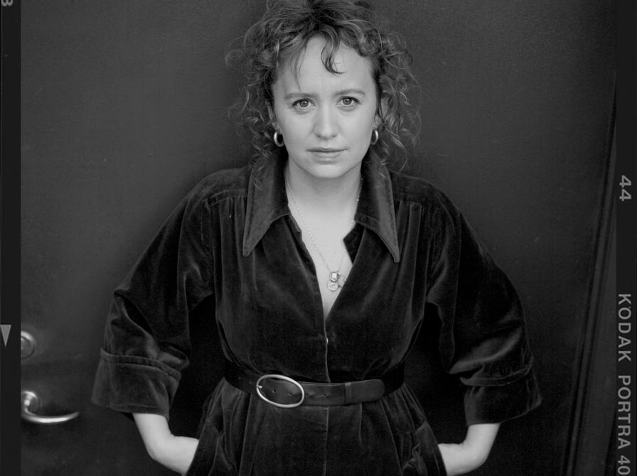 Sort-hvidt portræt af forfatteren Olga Ravn i mørk kjole med bælte, stående foran en mørk baggrund.